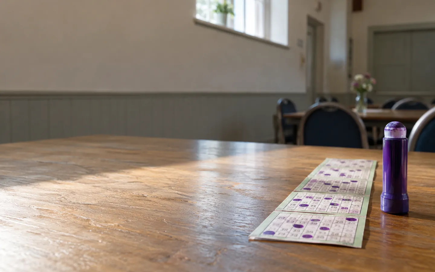 Paper 90-ball bingo tickets and a purple ink dabber on a table in a quiet community hall, with soft daylight from a high window